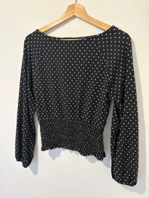 Svart prickig blus med smock - Snygg svart blus med vita små prickar och smockad midja. Blusen har långa ärmar med lätt puff och resår vid ärmslut. Tillverkad i ett mjukt, tunt tyg som ger ett avslappnat men trendigt intryck. Perfekt att matcha med jeans eller kjol. Storlek 158-164 men sitter som xs🤗