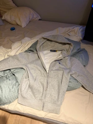 Ljusgrå hoodie med dragkedja - Säljer en stilren ljusgrå hoodie med dragkedja och huva. Hoodien har långa ärmar, ribbade muddar och fickor framtill. Perfekt för chill dagar eller när du vill ha en enkel och clean look. Materialet är mjukt och skönt mot huden. Säljer den pågrund av att den är för liten för mig.
