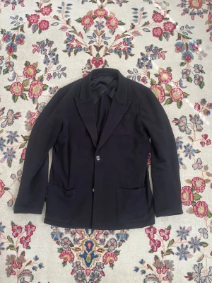 John Henric kavaj - Stilren svart kavaj med två knappar framtill, klassisk krage och tre fickor. Kavajen har en rak passform och är helfärgad utan mönster. Perfekt för dig som vill ha en enkel och tidlös look. Storlek M