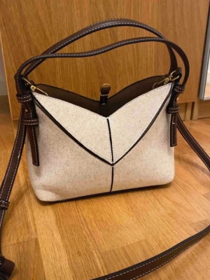 Beige axelväska med bruna detaljer - Snygg beige axelväska i canvas med bruna skinnremmar och dekorativa sömmar. Väskan har guldfärgade metalldetaljer och praktiska fästen för axelremmen. Perfekt storlek för vardagens essentials och stilren design.