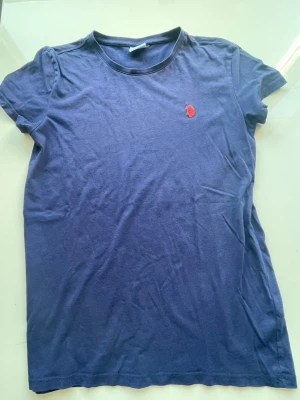 Blå t-shirt från U.S. Polo Assn - Mörkblå t-shirt från U.S. Polo Assn med klassisk passform och korta ärmar. T-shirten har en liten röd logga broderad på bröstet och är tillverkad i mjuk bomull. Perfekt till en avslappnad stil. Den är äkta.