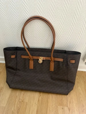 Brun Michael Kors handväska - Snygg brun handväska från Michael Kors med klassiskt MK-mönster och detaljer i ljusbrunt läder. Väskan har dubbla axelband och guldfärgat lås framtill med MK-logga. Perfekt storlek för att få plats med allt du behöver. Finns tyvärr en fläck på insidan! Pris går att diskutera vid snabb affär!
