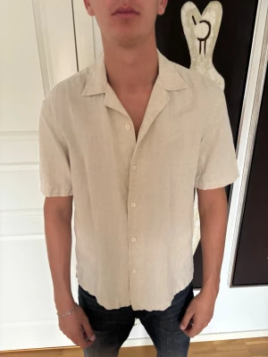 Beige linneskjorta med kort ärm - Säljer en stilren beige skjorta i linne med korta ärmar och klassisk krage. Skjortan har knappar framtill och en avslappnad passform, perfekt för varma dagar.