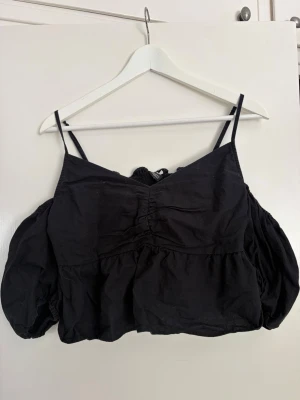 Svart croppad festtopp med puffärmar - Trendig svart croppad topp med tunna axelband och puffiga korta ärmar. Toppen har rynkade detaljer framtill och smockad rygg för extra komfort. Perfekt för dig som vill ha en cool och luftig look till sommaren. Storlek 40 men passar för mig som har S/M