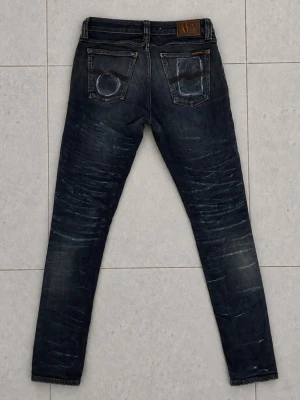 Nudie Jeans 30/34 - Säljer ett par mörkblå skinny jeans från Nudie Jeans med slitna detaljer och tvättade effekter på både fram- och baksida. Klassisk femficksmodell med läderpatch bak i midjan. Snyggt smal passform och coola slitningar för en trendig look.