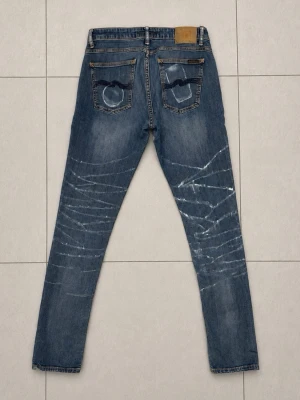 Nudie Jeans 31/34 - Säljer ett par blå jeans med unika vita slitningseffekter över benen och klassisk femficksdesign. Jeansen har raka ben och kontrastsömmar samt detaljerade bakfickor med broderi. Perfekta för dig som gillar en trendig och avslappnad stil.
