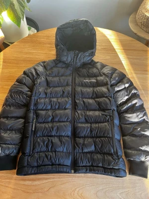 Svart dunjacka med huva - Snygg svart dunjacka med huva och dragkedja framtill. Jackan har två sidofickor med dragkedja och quiltad design som håller värmen bra. Perfekt för kalla vinterdagar och har en stilren look som passar till det mesta.