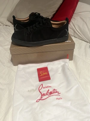 Svarta sneakers från Christian Louboutin - Snygga svarta låga sneakers från Christian Louboutin med klassisk röd detalj och signatur på hälen. Skorna har svart mocka, svarta skosnören och svart sula. Perfekta för dig som vill ha en stilren och exklusiv look. Sömnen vid sidan på ena skon har låsnat men inget svårt o fixa bara du har lim!