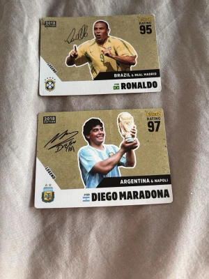 Fotbollssamlarbilder Maradona & Ronaldo/fotbollskort - Två samlarbilder från 2018 med fotbollslegenderna Diego Maradona och Ronaldo. Maradona håller i VM-pokalen och Ronaldo är i brasiliansk landslagströja. Båda korten har autograftryck och visar spelarnas rating.