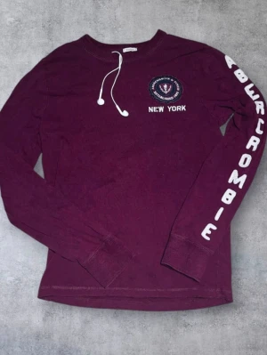 2000s ~Vinröd   Abercrombie & Fitch New York Long Sleeve Top - Snygg vinröd långärmad tröja från Abercrombie med tryck på bröstet och texten 'Abercrombie' längs ena ärmen. Rund halsringning och ribbade muddar. Perfekt för en avslappnad och trendig look.