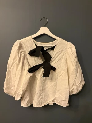 Vit blus med puffärmar och svarta rosetter - Trendig vit blus med stora puffärmar och två svarta rosetter i ryggen, som man knyter själv. Blusen har en lös passform och är tillverkad i ett lätt tyg som ger en luftig känsla. 