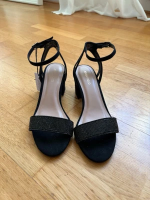 Svarta sandaler med klack från Anna Field - Stilrena svarta sandaler från Anna Field med bred rem framtill dekorerad med små svarta stenar. Skorna har en stadig blockklack och justerbar rem runt vristen. Perfekta för att lyfta din outfit med en touch av glitter. Aldrig använda!!! 
