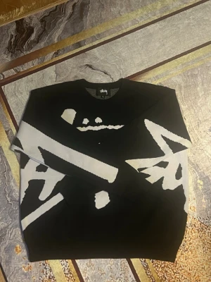 Svart stickad tröja från Stussy - Säljer en svart stickad tröja från Stussy med stort vitt grafiskt logotyptryck över hela framsidan. Tröjan har rund halsringning och ribbade muddar vid ärmslut och nederkant. Perfekt för dig som gillar streetwear och vill sticka ut.