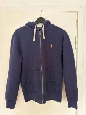 Polo ralph lauren zip hoodie  - Mörkblå hoodie från Polo Ralph Lauren med dragkedja framtill, vita snören i huvan och klassisk gul logga broderad på bröstet. Hoodien har kängurufickor och ribbade muddar vid ärmar och nederkant. Perfekt för en avslappnad och stilren look.