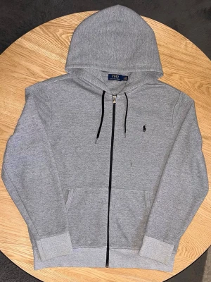 Polo Ralph Lauren Zip Hoodie Grå – Storlek L - Säljer en Polo Ralph Lauren zip-hoodie i grå melange.  Klassisk modell med svart polo-logo på bröstet, huva med svarta snören och känguruficka.  Storlek: L (LG)  Skick: Mycket bra. Tvättad och klar. Liten märke/fläck syns vid dragkedjan (se bild), annars fin.  Unisex.  Mått kan tas vid förfrågan. 