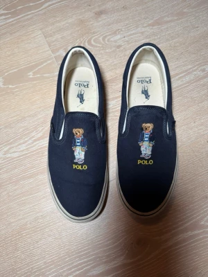 Marinblå slip-on sneakers Polo Bear - Marinblå slip-on sneakers från Polo Ralph Lauren med broderad Polo Bear på ovandelen och gul POLO-text. Skorna har vit sula och är tillverkade i canvasmaterial. Klassisk och stilren design med bekväm passform. Knappt använda, storlek 43