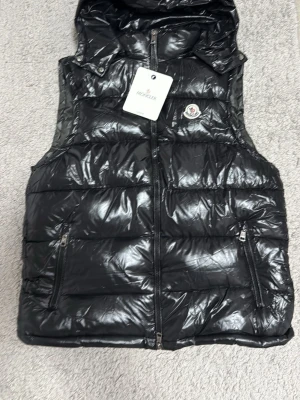 Svart dunväst från Moncler - Snygg svart dunväst från Moncler med glansig finish och huva. Västen har två dragkedjeförsedda fickor framtill och en klassisk Moncler-logga på bröstet. Perfekt för lager-på-lager och håller dig varm under kyliga dagar.
