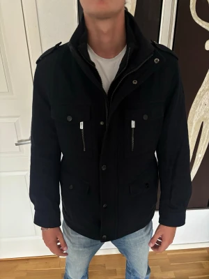 Jacka Michael Kors - Snygg svart parkajacka med hög krage, axelklaffar och flera fickor framtill. Jackan har dragkedjor och knappar samt förstärkta partier på armbågarna. Perfekt för dig som vill ha en stilren och funktionell jacka till vardags. Från Michael Kors med storlek M.
