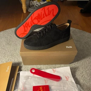 Svarta Louboutin sneakers med röd sula - Snygga svarta sneakers från Christian Louboutin med ikonisk röd sula och diskret logga undertill. Skorna har en klassisk låg siluett, svarta skosnören och är tillverkade i mocka för en lyxig känsla. Perfekta för dig som vill sticka ut med stil.