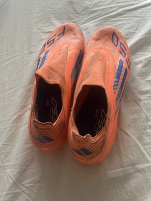 Orangea Adidas F50 fotbollsskor - Säljer ett par grymma orangea Adidas F50 fotbollsskor med blå detaljer och klassiska tre ränder på sidan. Skorna har en strömlinjeformad design utan snörning och är tillverkade i ett lätt syntetmaterial för maxad speed på planen.