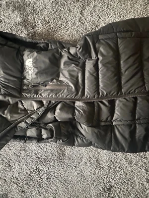 Svart dunväst från The North Face - Snygg svart dunväst från The North Face med ThermoBall-isolering. Västen har dragkedja framtill, en bröstficka med dragkedja och quiltad design. Perfekt för lager-på-lager och håller dig varm under kyliga dagar.