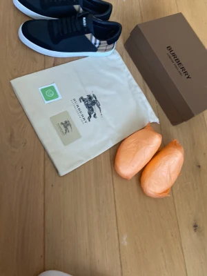 Burberry svarta sneakers med rutmönster - Snygga svarta sneakers från Burberry med klassiskt beige rutmönster på hälen och vita sulor. Skorna har svarta snören och detaljer i mocka och textil. Perfekta för dig som vill ha en stilren och exklusiv look. Ny pris 4000kr säljs för 2000kr pris kan diskuteras. Säljer de för att mina föräldrar köpte fel storlek