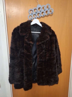 Mörkbrun fuskpälsjacka - Snygg mörkbrun fuskpälsjacka med bred krage och rak passform. Jackan har ett mjukt, fluffigt material och helsvart innerfoder. Perfekt för att hålla värmen under kalla dagar och ger en lyxig känsla till din outfit.
