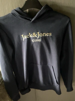 Mörkblå hoodie från Jack & Jones - Snygg mörkblå hoodie från Jack & Jones med en Klassisk känguruficka framtill och huva. Perfekt för dig som gillar enkel och stilren streetwear. Mjuk bomullskänsla och ribbade muddar vid ärmar och midja.