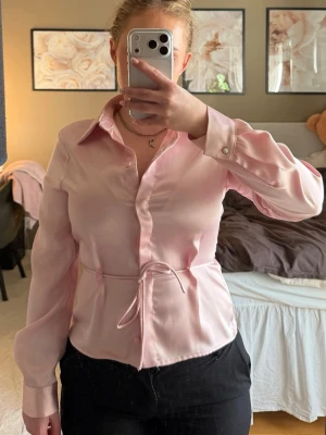 Ljusrosa satinskjorta med knytband - Säljer en ljusrosa skjortblus i satin. Blusen har klassisk krage, knappar framtill och ett smalt knytband i midjan för en snygg siluett. Långa ärmar med knapp vid manschetten. Perfekt för en chic och stilren look.