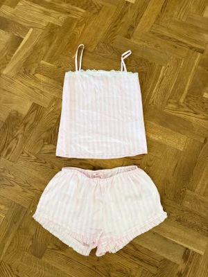 Rosa randig pyjamas med spetsdetalj - Söt pyjamas i två delar med ljusrosa och vita ränder. Linnet har tunna axelband och spetskant upptill, medan shortsen har volangkant nedtill. Perfekt för mysiga kvällar hemma. Materialet känns mjukt och lätt mot huden. Små defekter förekommer men inget märkvärdigt. Storlek 158/164 motsvarande XS.
