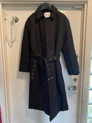 Svart trenchcoat med bälte - Stilren svart trenchcoat med klassisk krage och långt snitt. Jackan har ett matchande bälte med metallspänne och flera hål för justering. Långa ärmar och knäppning framtill. Perfekt för dig som gillar minimalistisk och tidlös stil.