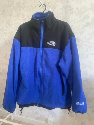 Blå fleecejacka från The North Face - Blå och svart fleecejacka från The North Face med dragkedja framtill, hög krage och två sidofickor. Jackan har Windstopper-tyg på ärmen och logga på bröstet och ryggen. Perfekt för kyliga dagar och outdoor-äventyr.