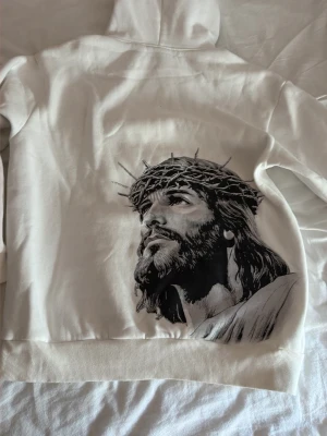 Vit hoodie med Jesusmotiv och kors - Säljer en vit hoodie med stor svartvit illustration av Jesus med törnekrona på ryggen och ett litet kors på bröstet. Hoodien har huva med dragsko och magficka. Tillverkad i mjukt material, perfekt för dig som gillar unika tryck och vill sticka ut. Passar både kvinna och man storlek M, den här en liten fläck. 