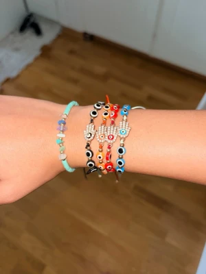 Färgglada armband med pärlor och ögon - Säljer flera armband med färgglada pärlor och ögonmotiv. Några har hamsa-händer dekorerade med små stenar. Armbanden har olika färger som svart, orange, röd, blå och turkos. Perfekta för dig som gillar unika accessoarer och vill sticka ut.