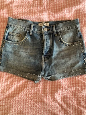 Blå jeansshorts med nitar Pull&Bear - Säljer ett par blå jeansshorts från Pull&Bear med coola nitar längs fickor och benslut. Shortsen har råa kanter och klassisk femficksdesign. Perfekta för sommaren och ger en edgy vibe till din outfit. (Sytt om ett par jeans till dessa shorts!)