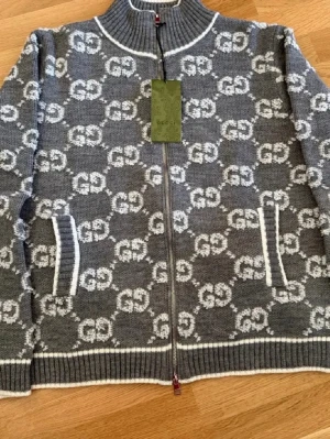 Grå stickad ziptröja från Gucci - Snygg grå stickad ziptröja från Gucci med vitt GG-mönster över hela plagget. Tröjan har hög krage, långa ärmar och ribbade muddar samt vita detaljer vid fickor och kanter. Perfekt för dig som vill ha en trendig och exklusiv look.