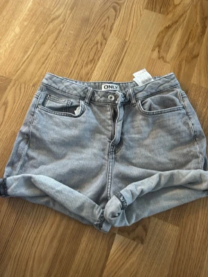 Grå denimshorts från ONLY - Säljer ett par grå denimshorts från ONLY med hög midja och upprullade benslut. Klassisk femficksmodell med knapp och dragkedja framtill. Perfekta för sommaren och lätta att matcha med olika toppar.
