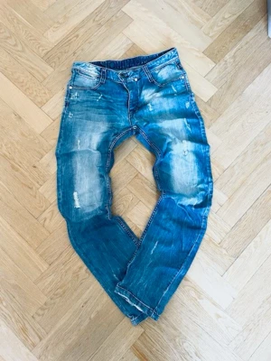 Blå slitna jeans med tvättade detaljer Humör  - Säljer ett par blå jeans med slitna och tvättade detaljer, raka ben och klassisk femficksmodell. Jeansen har ljusa partier och små revor för en trendig, avslappnad look. Perfekta för dig som gillar en casual streetstil. Modell Jalle. Innerbenslängd 85cm har ett hål vid fickan