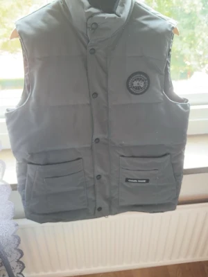 Grå väst från Canada Goose - Säljer en grå väst från Canada Goose. Västen har ett märke på bröstet och knäppning med svarta knappar. Perfekt för att hålla värmen under kyliga dagar.