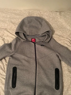 Grå Nike hoodie med dragkedja XL - Snygg grå hoodie från Nike i storlek XL med hel dragkedja framtill och två svarta dragkedjeförsedda fickor. Klassisk Nike-logga på bröstet och stor huva. Perfekt för chill eller träning. Tillverkad i mjukt material för extra komfort.