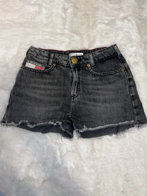 Mörkgråa jeansshorts med rå kant - Säljer ett par mörkgråa jeansshorts med rå, fransig kant och klassisk femficksdesign. Shortsen har knappgylf och bälteshällor. De är tillverkade i denim. Midwaist.