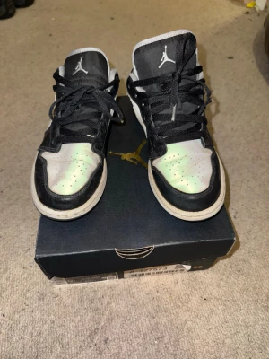Svarta och vita Air Jordan 1 Low - Säljer ett par Air Jordan 1 Low sneakers i svart och vitt med grå Swoosh. Klassisk siluett med perforerad tåbox, svarta skosnören och vit sula. Ikonisk Jumpman-logga på plösen och stilren design som passar till streetwear.