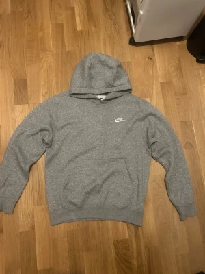 Grå hoodie från Nike - Klassisk grå hoodie från Nike med vit logga på bröstet. Hoodien har en stor ficka framtill, ribbade muddar vid ärmar och nederkant samt en rymlig huva. Perfekt för en avslappnad och sportig stil.