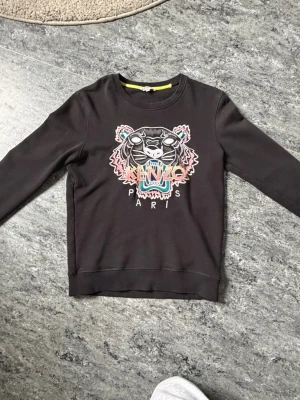 Svart sweatshirt från Kenzo med tiger - Cool svart sweatshirt från Kenzo med ikoniskt tigerhuvud och färgglad brodyr på bröstet. Tröjan har rund halsringning och långa ärmar. Perfekt för dig som vill sticka ut med en statement-piece från ett välkänt märke.