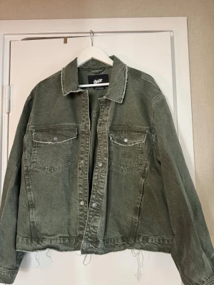 Grön oversized jeansjacka - Snygg grön jeansjacka i oversized modell med råa kanter vid krage och nederkant. Två stora bröstfickor med knappstängning och klassisk knappslå framtill. Perfekt för dig som gillar en avslappnad och trendig stil.