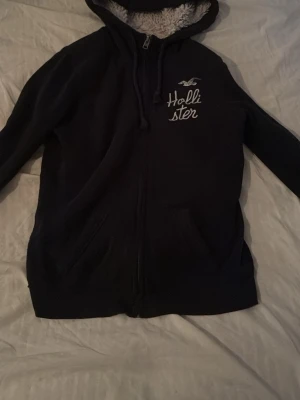 Svart hoodie från Hollister med dragkedja - Mysig svart hoodie från Hollister med vit broderad logga på bröstet och dragkedja framtill. Hoodien har en fluffig, vit fuskpälsfodrad huva och två fickor fram. Perfekt för chill dagar och enkel att matcha med jeans eller mjukis. Priset kan diskuteras 
