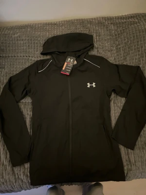 Svart windbreaker från Under Armour - Snygg svart hoodie från Under Armour med dragkedja framtill och vit logga på bröstet. Stor huva och vita detaljer vid axlarna. På ryggen står det 'UNDER ARMOUR' i vitt och ärmen har 'STORM'-tryck. Tillverkad i ett tekniskt material som står emot väta.