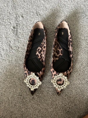Leopardmönstrade ballerinaskor Zara - Säljer ett par ballerinaskor från Zara med leopardmönster och spetsig tå. Skorna har en stor glittrig dekoration framtill med gnistrande stenar. Materialet känns som satin och färgerna går i beige och brunt. Perfekta för dig som vill sticka ut!