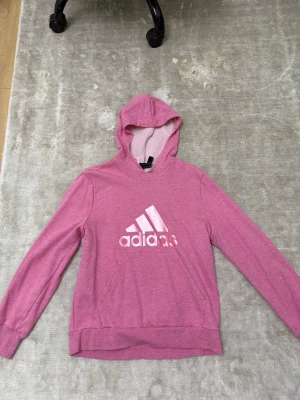 Rosa hoodie från Adidas - Rosa hoodie från Adidas med klassisk logga framtill. Tröjan har huva, långa ärmar och en stor magficka. Perfekt för en avslappnad och sportig stil. Mjuk bomullsblandning som känns skön mot huden.