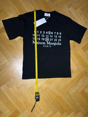 Maison Margiela svart t-shirt - Svart t-shirt från Margiela med ikoniskt tryck framtill i vitt med siffror och logga. Klassisk passform och rund hals. Perfekt för dig som gillar stilrena och trendiga plagg med designer-touch. Fick i present vet ej om real